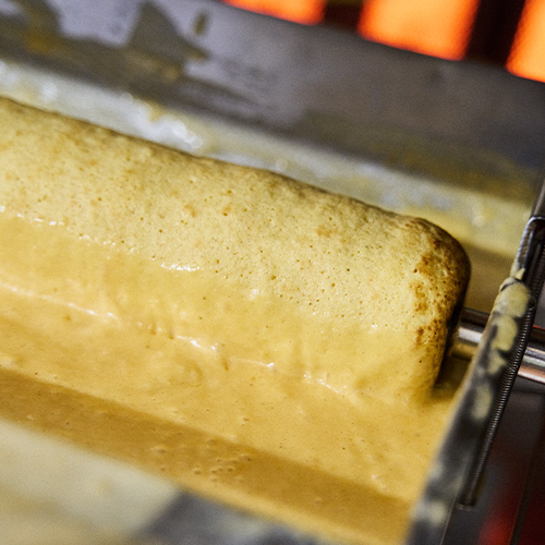Auf Spiess drehender, goldbrauner Teig wird ueber heisser Flamme gebacken.