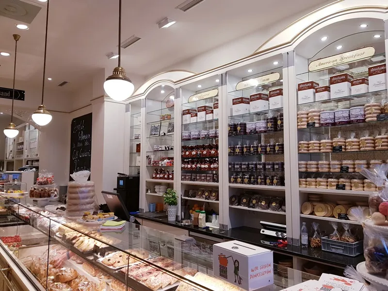 Konditorei mit Gebaeckvitrine. Verkaufsregale gefuellt mit Marmeladen- und Keksdosen. Im Hinter- grund steht "American Breakfast".

Text: 
Wir sagen Dankeschoen