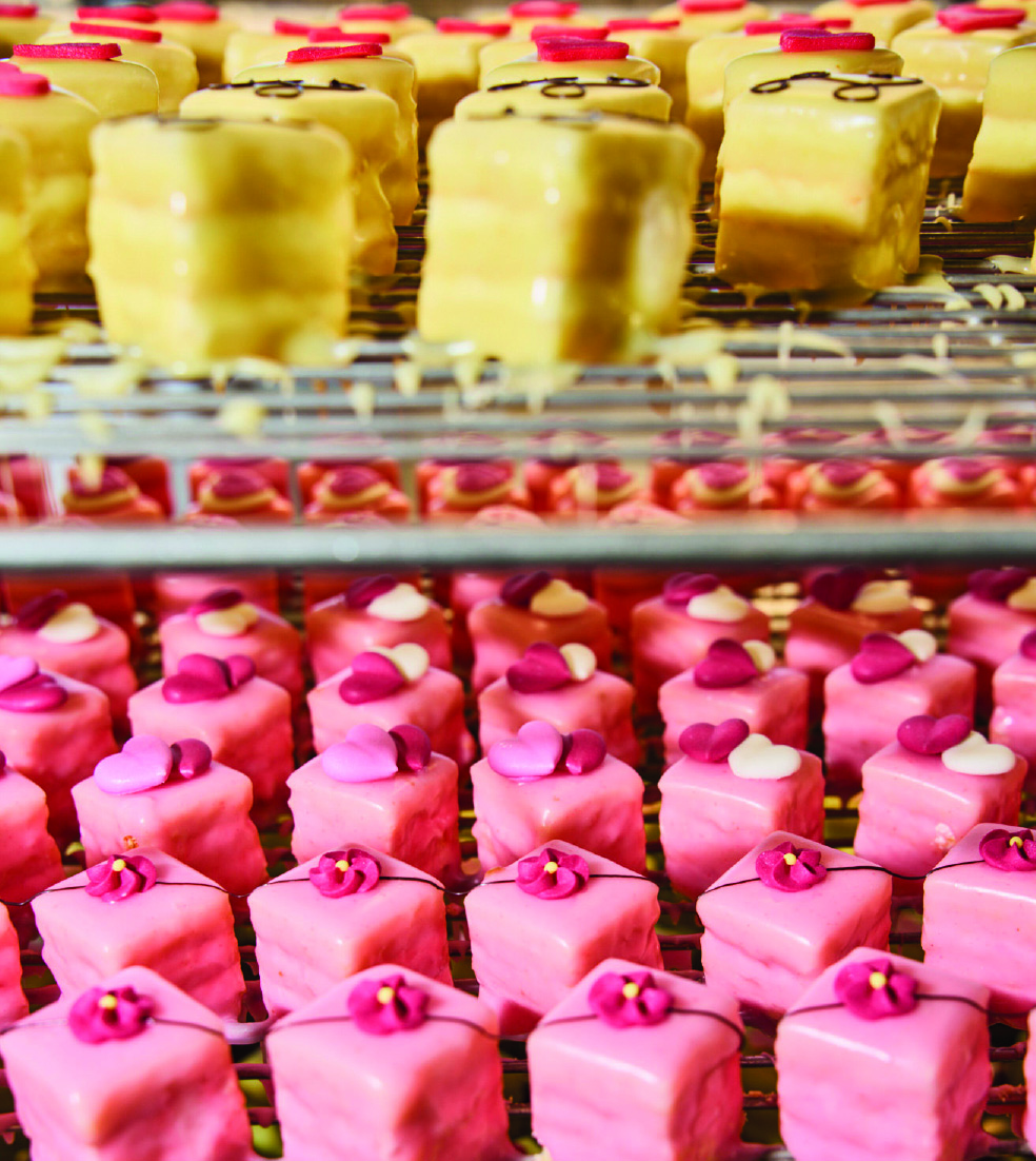 Mehrere Reihen kleiner gelber und rosa Gebaeckwuerfel auf Gittern, verziert mit bunten Blumen.